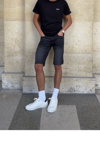 Homme portant un t-shirt noir, un short en denim foncé, des chaussettes blanches et des baskets blanches, debout les mains dans les poches contre un mur en pierre.