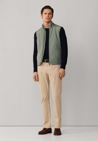 Mann trägt eine hellgrüne ärmellose Weste über einem dunkelblauen Pullover, beigefarbene Hose, geflochtener brauner Gürtel und braune Loafers, steht vor einer schlichten Wand.