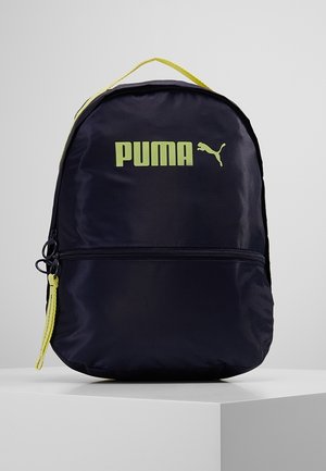 Mochila negra Puma con asa amarilla, bolsillo frontal con cremallera y logo amarillo de Puma sobre una superficie blanca de exhibición.