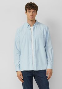 Chemise bleu clair en coton avec une poche poitrine, un col et des manches longues. Modèle debout portant un jean foncé.