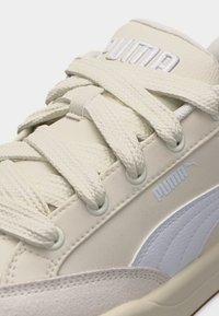 Κοντινή λήψη μπεζ αθλητικού παπουτσιού Puma με χοντρές κορδόνια, κεντημένο λογότυπο και λευκή πλευρική επιφάνεια με υφή.