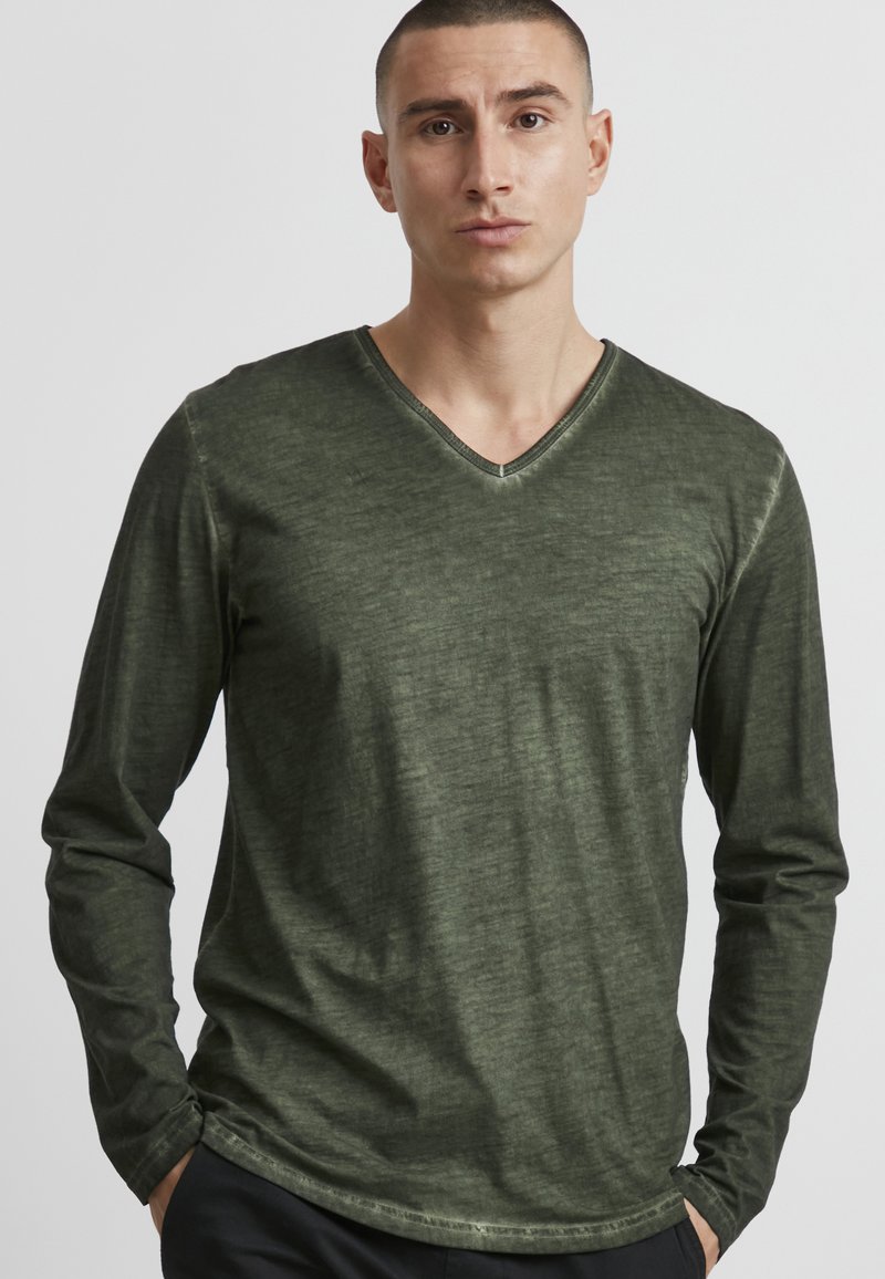 Langärmliges V-Ausschnitt-Shirt in dunkelolivgrün, aus weichem Material mit subtiler Textur und kontrastierenden Nähten entlang des Ausschnitts.