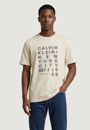 Jeune homme portant un T-shirt beige Calvin Klein avec un texte vertical faisant référence à New York City et 1968, associé à un jean bleu.