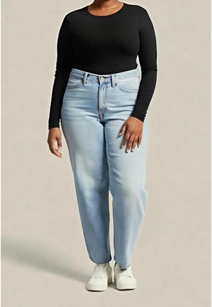 Personne portant un jean taille haute bleu clair, un haut noir à manches longues et des baskets blanches, debout devant un fond uni.