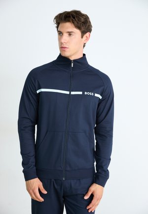 Marineblå zip-up treningsjakke laget av elastisk stoff, med en hvit horisontal stripe, stående krage og frontlommer.