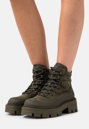 Jambes portant des bottes de randonnée en cuir suédé vert olive avec des semelles épaisses et crantées et un laçage à l'avant sur un fond blanc.