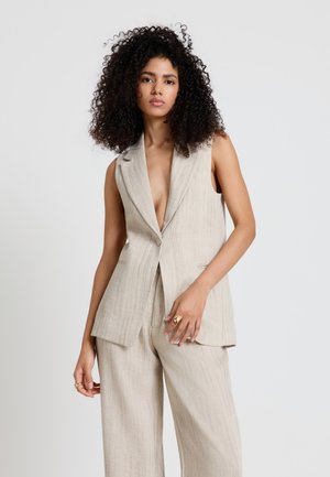 Frau mit lockigem Haar, die einen beigen ärmellosen Blazer und passende weite Hosen trägt, steht vor einem einfarbigen hellen Hintergrund.