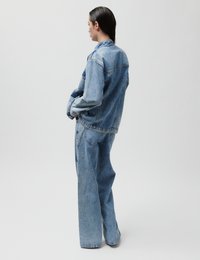 Γυναίκα με φαρδιά, ανοιχτό μπλε τζιν μπουφάν και τζιν παντελόνι με φαρδιά μπατζάκια στέκεται πλαγιαστά μπροστά σε απλό γκρι φόντο.