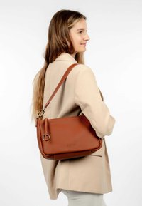 Femme en blazer beige portant un sac à bandoulière en cuir tan avec des accents en métal argenté, regardant à droite sur fond blanc.
