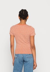 Halenka s krátkým rukávem, přiléhavá, v měděné barvě s bílým květinovým potiskem, hladká textura, kulatý výstřih a jednoduchý pohled zezadu. V kombinaci se světle modrými džíny.