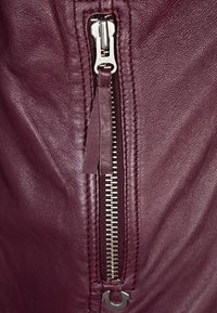 Giacca di pelle borgogna con zip metallica dotata di un tirante in tessuto, cuciture a vista e denti della zip in contrasto.