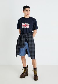 Navy zilā kokvilnas T-krekls ar nodilušu Union Jack dizainu, apvienots ar džinsu šortiem un melnu raibu krekli, kas sasaistīts ap vidu; brūni zābaki.