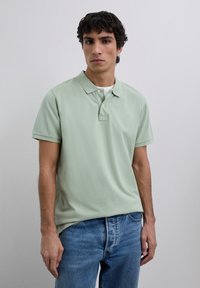 Nuori mies, joka pukeutunut vaaleanvihreään lyhythihaiseen polo-paitaan valkoisen t-paidan päällä ja sinisiin farkkuihin, seisoo yksinkertaista vaaleanharmaata taustaa vasten.