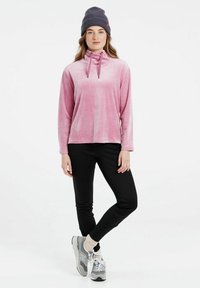 Pullover lungo rosa in velluto con collo alto e cordini, abbinato a pantaloni neri e sneakers grigie. Presenta un berretto lavorato a maglia.