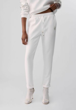 Persona que lleva pantalones jogger blancos con cordón, combinados con calcetines blancos y tacones abiertos, de pie contra un fondo liso.