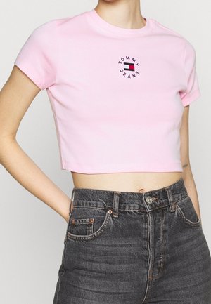 Kobieta nosząca różowy krótki t-shirt z logo Tommy Jeans i ciemnoszare jeansy z wysokim stanem, ręka oparta na biodrze.