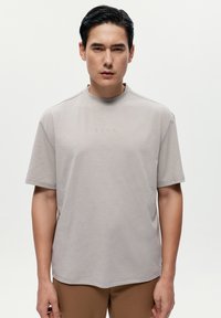 Světle šedá tričko s krátkým rukávem a kulatým výstřihem, které má minimální texturu a decentní branding přes oblast hrudníku.