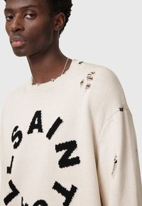 Cremfarbener Strickpullover mit schwarzen, abgenutzten Buchstaben. Verfügbar mit einem runden Ausschnitt, fransigen Kanten und mehreren kleinen Löchern für einen strukturierten Look.