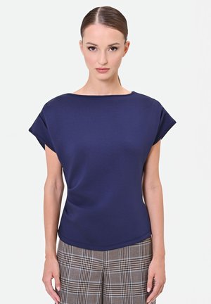 Donna con capelli tirati indietro che indossa una maglietta blu navy a maniche corte e pantaloni marroni a quadri, in piedi davanti a uno sfondo bianco uniforme.