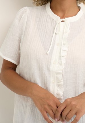 Blouse blanche texturée à manches courtes, avec un avant à boutons et une bordure en dentelle froncée. Présente un nœud décoratif au niveau du col.
