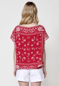 Top rosso a maniche corte con un motivo paisley bianco. Vestibilità ampia con scollo largo e dettagli decorativi lungo l'orlo e le maniche.