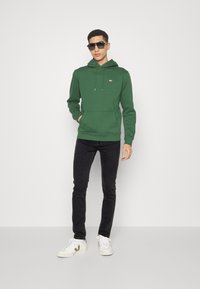 Sudadera verde con un bolsillo delantero, capucha con cordón y un pequeño logo en el pecho, combinada con jeans negros y zapatillas blancas.