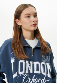 Sudadera de navy con media cremallera, que presenta el texto y gráficos en blanco de "LONDRES". Tela suave con un corte relajado y puños acanalados.