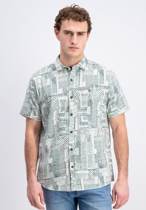 Man in een kortärmelig overhemd met abstracte zwarte en groene geometrische patronen, gecombineerd met een blauwe spijkerbroek, staand met een neutrale uitdrukking.