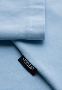 Tissu bleu clair avec coutures visibles et une étiquette noire de vêtement portant la mention « LEVEL UP » cousue sur le bord.