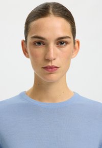 Jeune femme aux cheveux plaqués en arrière, portant un pull bleu clair, regardant droit devant avec une expression neutre sur fond blanc.