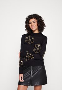Schwarzer Pullover mit hohem Kragen, der mit goldenen Schneeflocken-Designs verziert ist. Genutzt zusammen mit einem schwarzen Lederrock mit einem Reißverschlussdetail vorne.