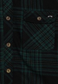 Green and black plaid flannel shirt featuring a button-up front, brown buttons, and a left chest pocket with a logo tag.