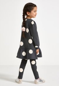 Donkergrijze jurk met grote crèmekleurige stippen, ruchezoom, gecombineerd met bijpassende leggings. Vlecht haar met een bloemenaccessoire. Zilveren schoenen.