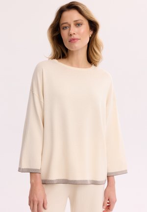 Kobieta ubrana w luźny kremowy top z długim rękawem i pasujące spodnie, z delikatnym paskowanym obszyciem na mankietach i u dołu, stojąca na jasnym tle.