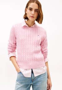 Hellrosa Pullover mit Zopfmuster und geripptem Saum, getragen über einem weiß-rosa gestreiften Hemd mit Kragen, kombiniert mit hellblauen Denim-Jeans.