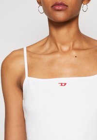 Hvit tanktopp med tynne stropper, med en liten rød brodert logo i midten. Myk, ribbet tekstur og firkantet utringning.