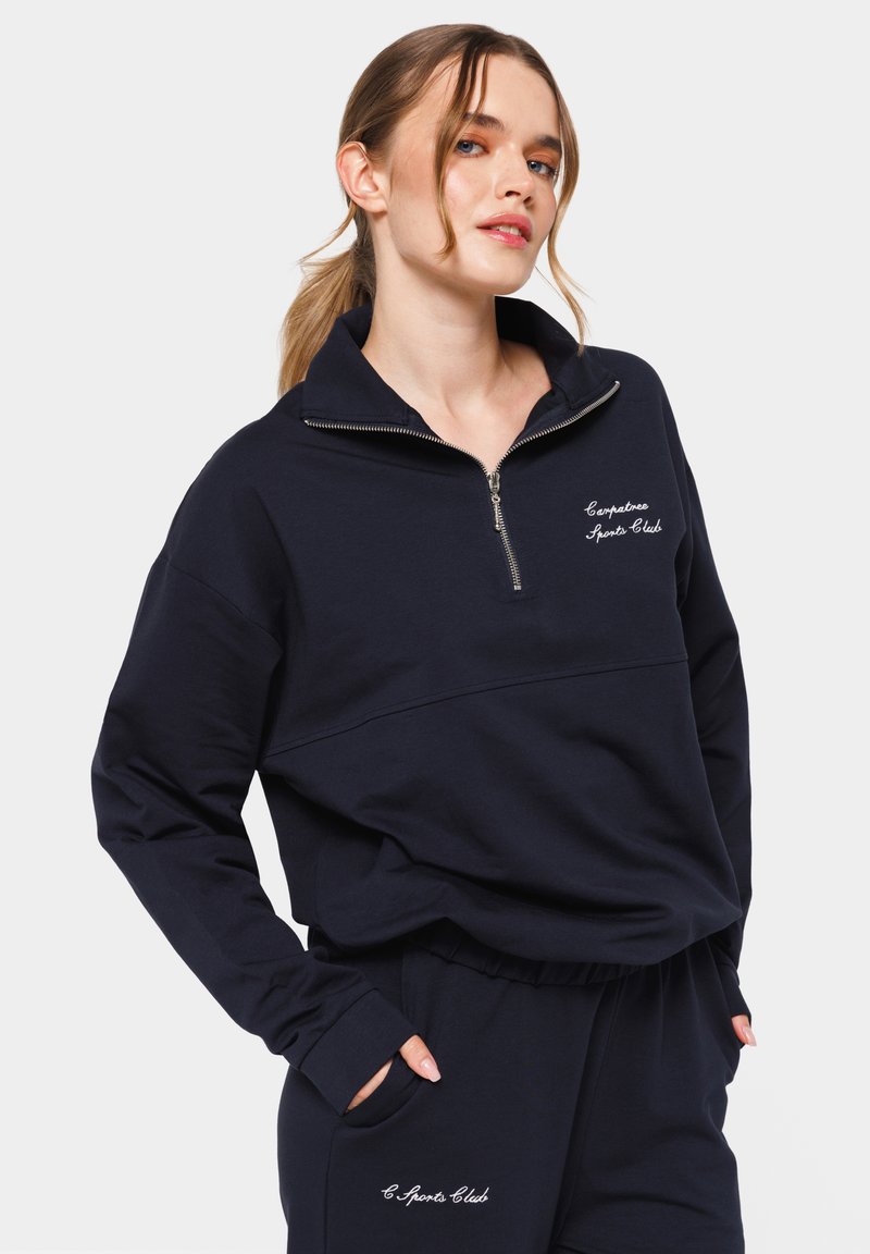 Sweatshirt marine avec col zippé, en mélange de coton doux, présentant un texte brodé blanc "C Sports Club" sur la poitrine et les poches.