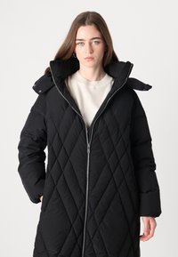 Crni puffer kaput s kvadratnom piktroskom sa punim zatvaračem, visokim ovratnikom i odvojivom kapuljačom. Ima dijamantni uzorak i bočne džepove.