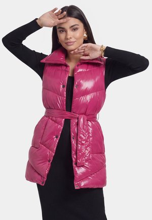 Frau trägt eine glänzende pinke gesteppte Weste mit Gürtel über einem schwarzen langärmeligen Kleid, posiert mit den Händen nahe am Gesicht und trägt goldene Ohrringe und Armband.