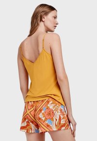 Top amarillo sin mangas con tirantes finos, combinado con pantalones cortos estampados que presentan diseños de flores y pájaros en colores naranja, azul y crema.