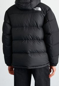 Chaqueta de plumas negra con capucha, presenta secciones acolchadas horizontales y un logo bordado en blanco en el hombro. Material suave e insulated.