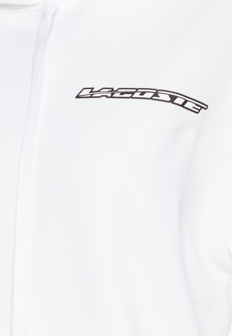 Biała bluza z kapturem z eleganckim czarnym logo "Lacoste". Wykonana z miękkiego materiału o gładkiej fakturze, z zamkiem błyskawicznym z przodu.
