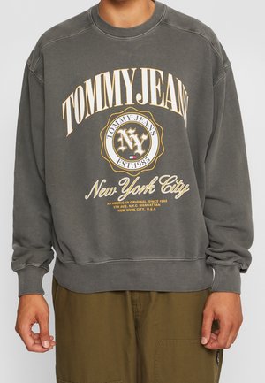 Osoba nosząca szarą bluzę z napisem i logo „Tommy Jeans New York City”, w połączeniu z oliwkowozielonymi spodniami.