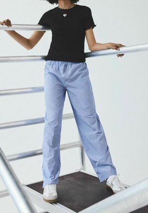 Haut noir à manches courtes avec bords festonnés, associé à un pantalon ample rayé bleu clair. Mannequin portant des baskets blanches. Rampe en métal en arrière-plan.