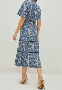 Sinivalkoinen paisley-kuvioinen mekko, jossa on lyhyet hihat, vyötärömuotoilu, liehuva helma ja mustat pitsikoristeet. Yhdistetty kultakorkoisiin sandaaleihin.