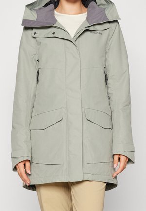 Lysegrøn vinterjakke med hætte fra Didriksons med store forlommer, lynlås og trykknapslukning, båret over beige bukser.