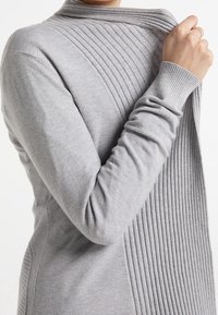 Light gray sweater with a ribbed texture on one side and smooth fabric on the other, featuring a high collar and fitted sleeves.