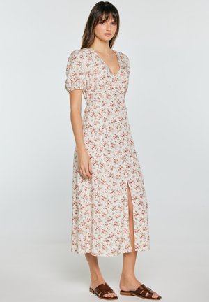 Kobieta stojąca w białej sukience midi z małym czerwonym kwiatowym nadrukiem, bufkami, dekoltem w serek, przednim rozcięciem i brązowymi płaskimi sandałami typu slide.