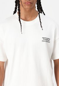 Witte katoen T-shirt met een ronde halslijn en korte mouwen. Bevat een kleine zwarte print op het linkerdeel van de borst. Soepele textuur.