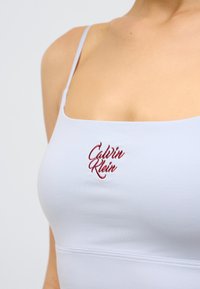 Ανοιχτό μπλε crop top με λεπτές τιράντες, με κεντημένο λογότυπο "Calvin Klein" σε σκούρο κόκκινο στο μπροστινό μέρος. Λείο, ελαστικό ύφασμα.